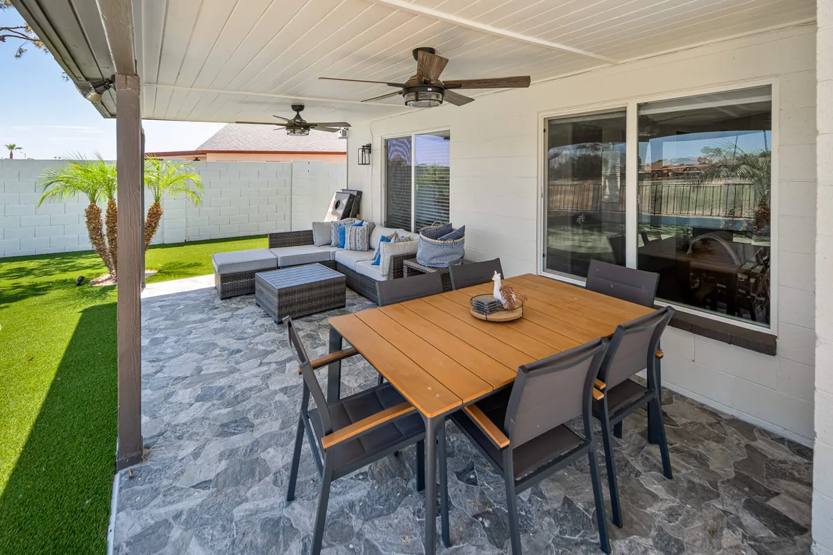 This exterior view showcases a beautifully furnished outdoor dining and lounge area set amidst palm trees. With comfortable seating, ceiling fans, and a backdrop of blue sky, it offers a glimpse of the property's inviting Southwestern-style retreat for post-golf relaxation.