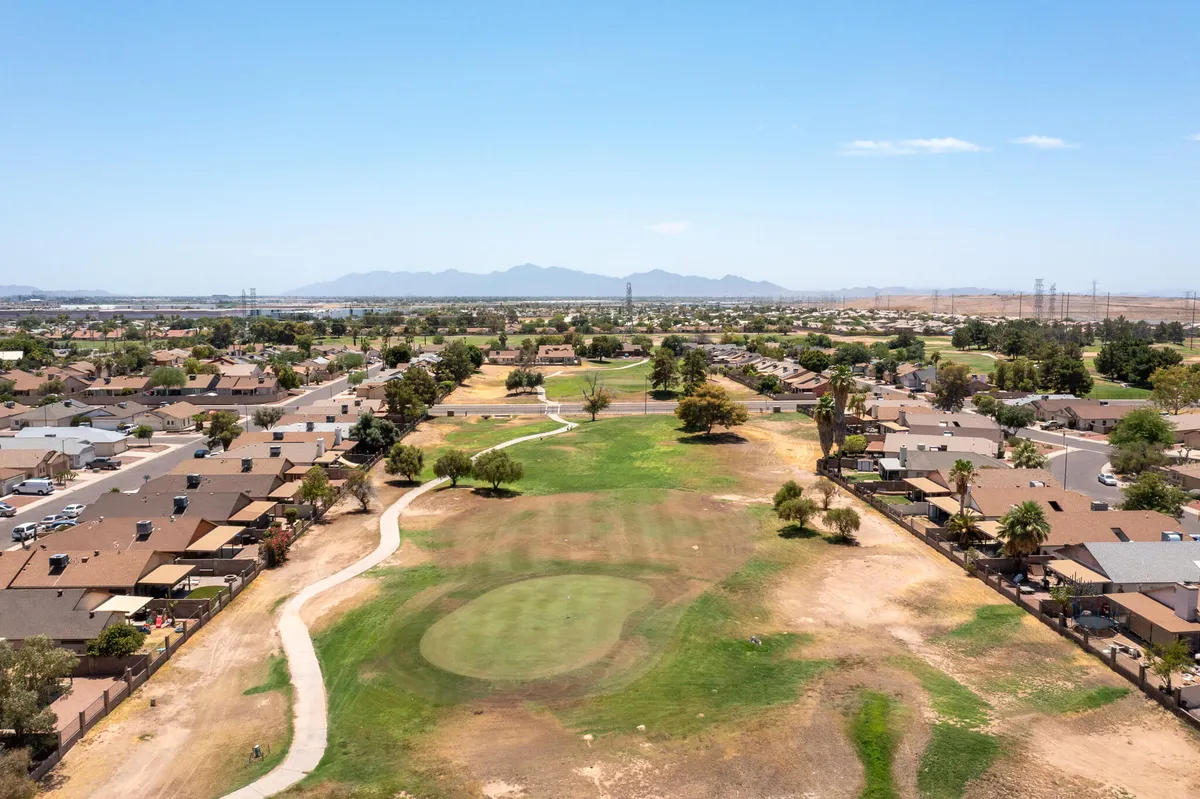 A wide-angle view of a lush golf course bordered by residential houses and backed by a dramatic mountain range. The scene is tranquil, with open skies and expansive green fairways, evoking the appeal of nearby courses mentioned in the section.