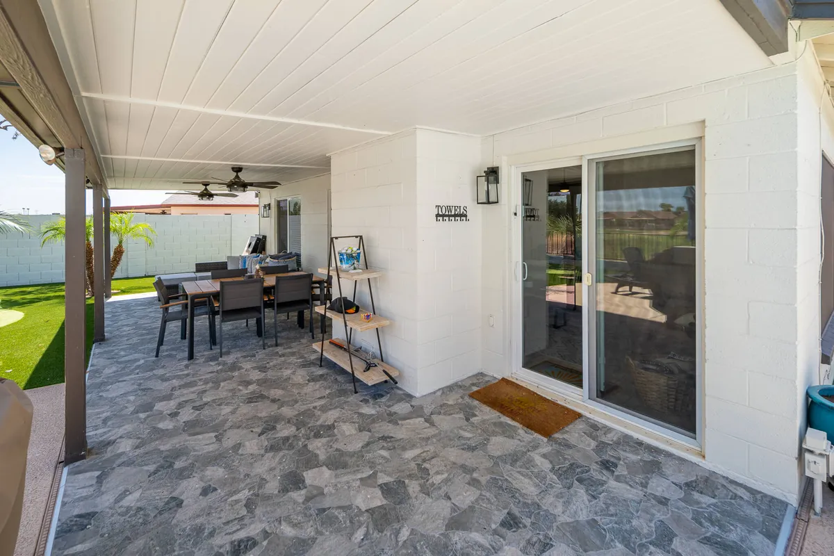 This image features an outdoor dining table and seating area on a patio, with a potted palm tree and a sliding glass door in the background. The scene is sunlit and inviting, ideal for gathering or relaxing after attending a local game or event.