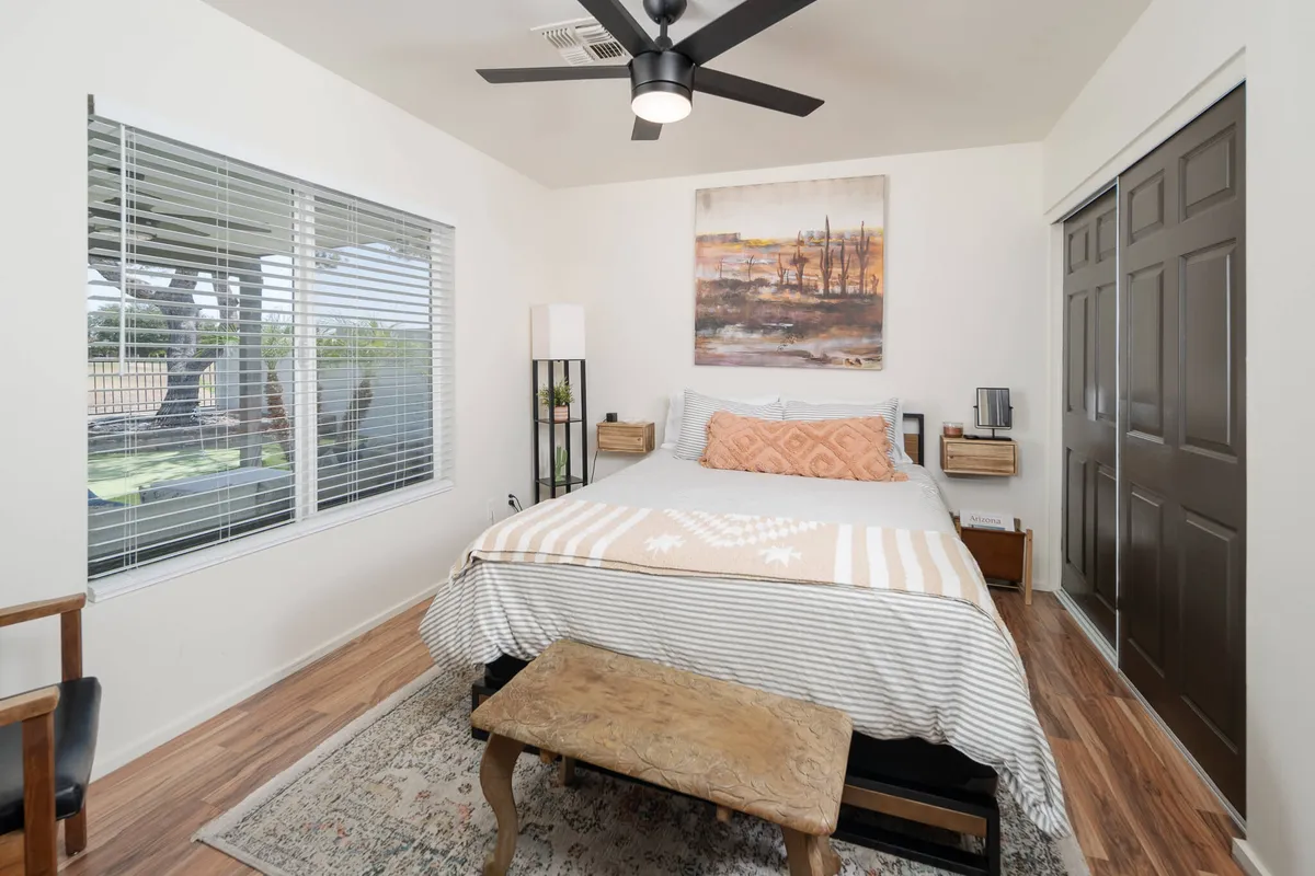 This image showcases a serene bedroom featuring a bed with decorative pillows, artwork above the bed, a nightstand with a lamp, and a window with blinds gently letting in natural light. The room’s palette and accents evoke a Southwestern calm, creating a sense of restful privacy.