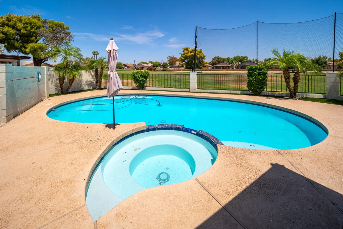 A sparkling swimming pool and jacuzzi surrounded by lush greenery and shaded by an umbrella. The inviting pool deck is perfect for swimming, sunbathing, or relaxing, with ample space for family fun under the Arizona sun.