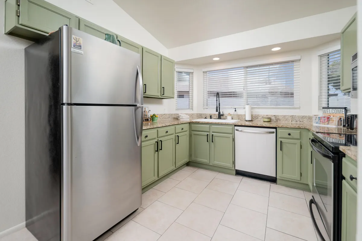 A bright and inviting kitchen featuring modern stainless steel appliances, green cabinetry, expansive granite countertops, and a clean sink area under large windows that bathe the space in natural light. The layout encourages gathering, with the kitchen island and ample counter space ready for meal prep or casual conversation.