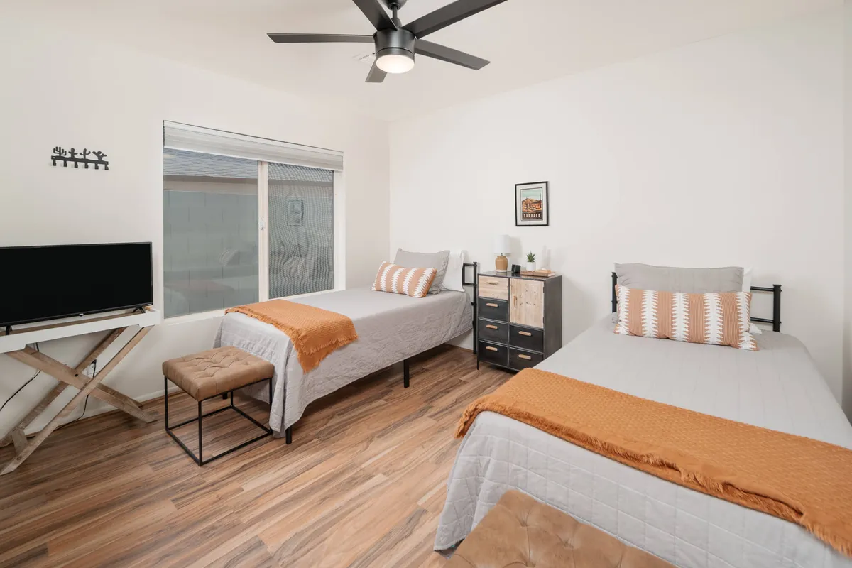 A cozy twin-bedroom setup with two beds, a television set, decorative wall art, a wooden nightstand, and a ceiling fan. The layout invites shared moments or casual relaxation, with details that suggest both comfort and thoughtful design.