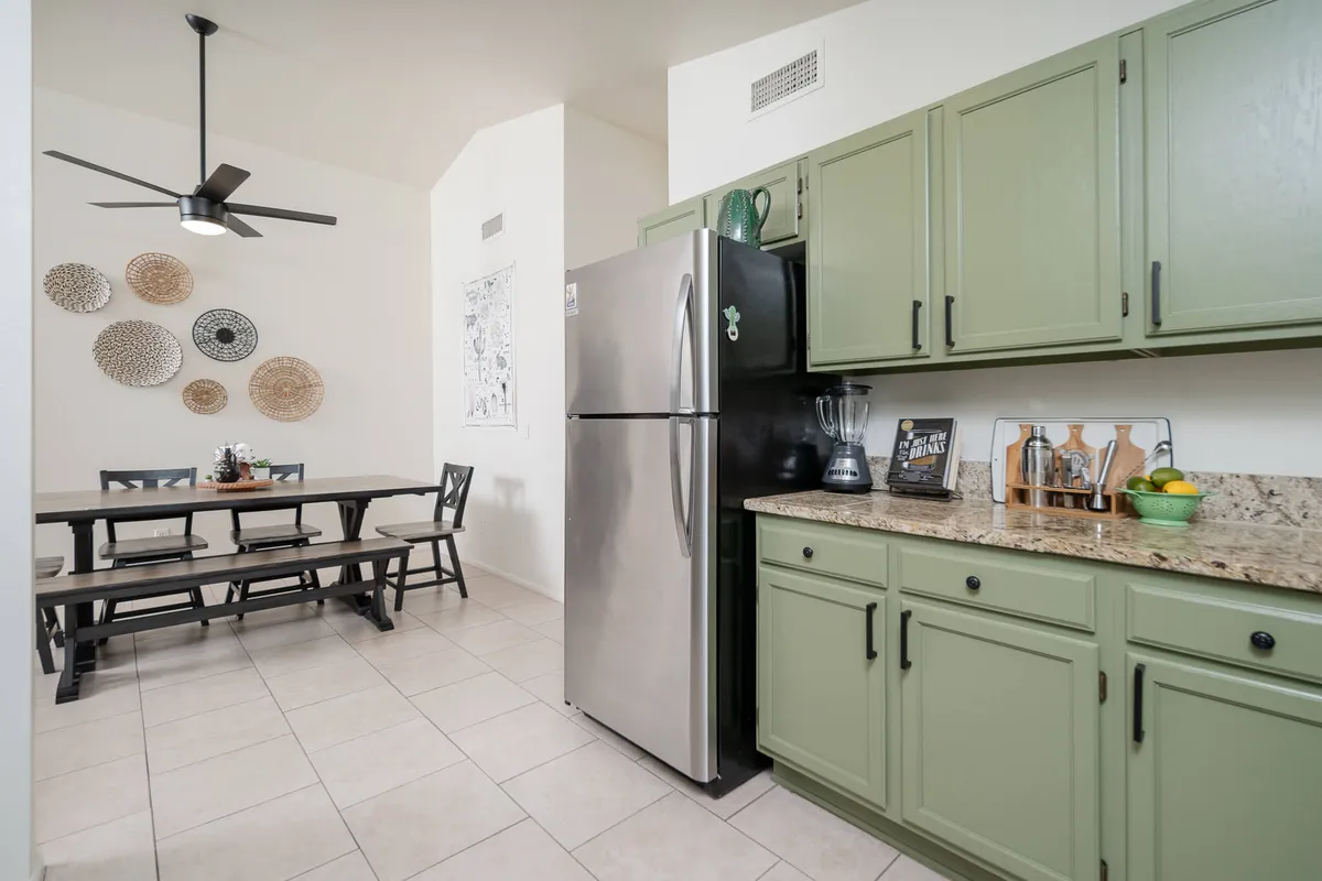 A cozy kitchen-dining area featuring green kitchen cabinets, a modern dining table, decorative wall plates, and stainless steel appliances. The set-up is warm and inviting, ideal for both meal preparation and sharing food with guests.